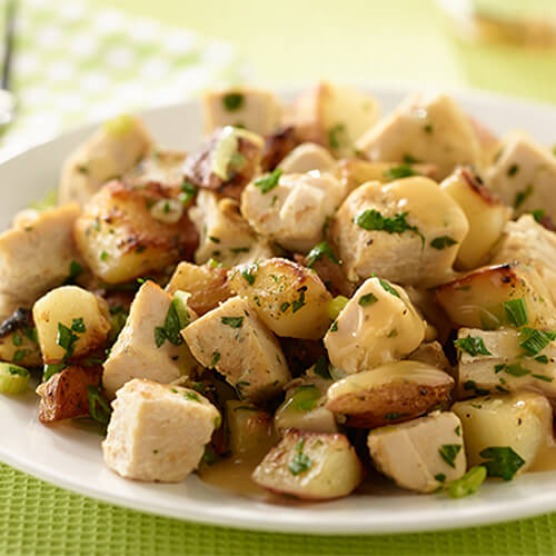 Cooked potatoes and onion, topped with salt and pepper, stirred with green onions and parsley, topped with gravy on a white plate atop a green tablecloth.