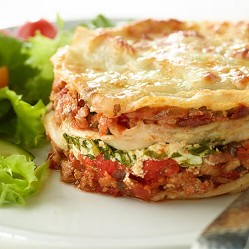 A cupcake-sized lasagna filled with ground turkey, cheese, tomato sauce, and vegetables, served on a white plate with a side of salad.