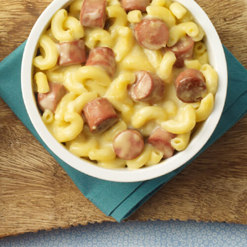 A fair portion of macaroni and cheese, topped and filled with cut turkey franks, served on a white plate, on a cyan napkin, atop a wooden table.