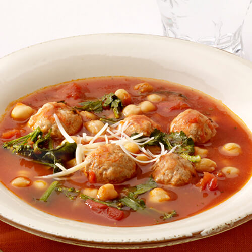 Ground turkey meatballs simmered in a savory tomato broth with fresh chickpeas and rapini., served in a white bowl and on an orange napkin.