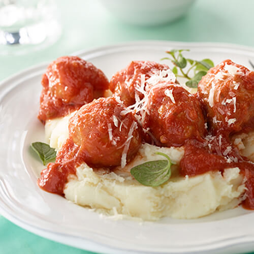 A hearty portion of marinara-topped meatballs atop a great portion of mashed potatoes in a white plate, on a turquoise tablecloth.