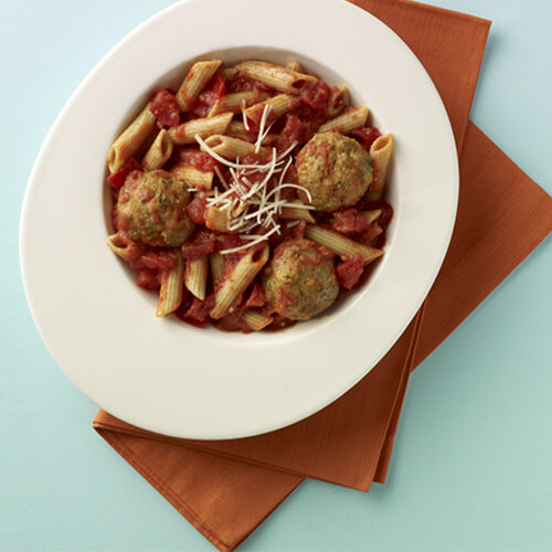 A great portion of penne pasta, covered in tomato sauce, and topped with meatballs made from turkey, garbanzo beans, and spices, served with parmesan cheese, in a white bowl on a red napkin.