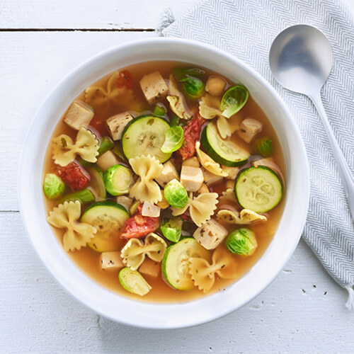 A savory broth filled with bowtie pasta, a hearty amount of vegetables, and turkey, served on a white plate.