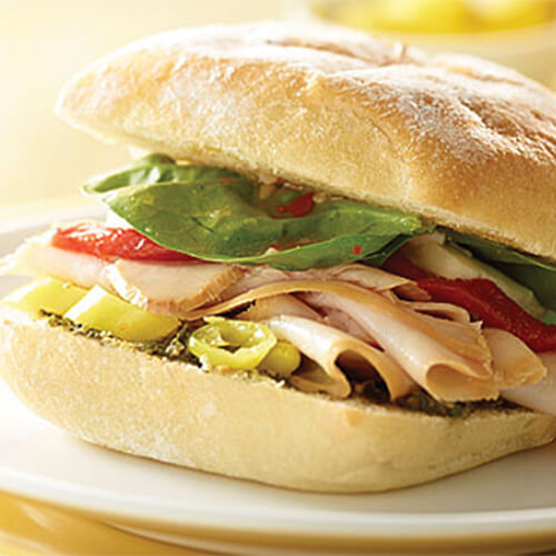 Spinach leaves, peppers, pesto, and smoked turkey, served on a cibatta roll, with a side of olives on a white plate atop a yellow table.