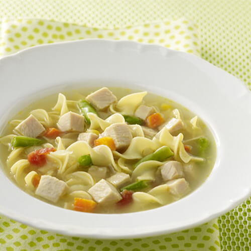 A healthy portion of noodles, turkey breast, chicken bouillon and vegetables in a white bowl on a green napkin and tablecloth with a green spoon.