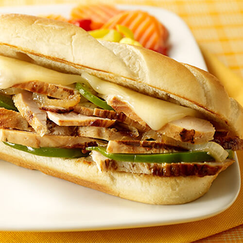 A hearty portion of Provolone, turkey strips, and bell peppers, served on a baguette on a white plate with a side of fruit atop a yellow cloth.