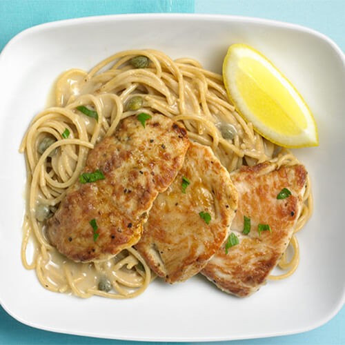 Angel hair pasta covered in a light wine sauce, topped with turkey tenderloin is gently simmered with lemon pepper and garnished with parsley and a lemon wedge, served on a white plate, and a teal tablecloth.