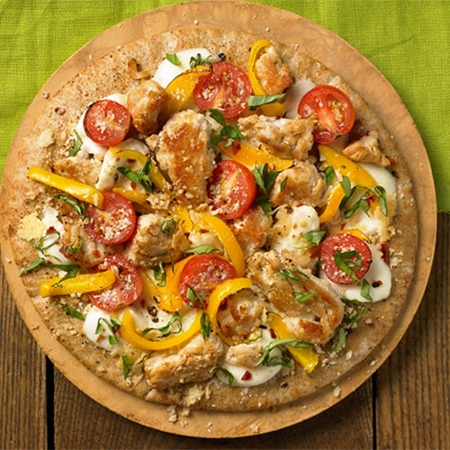 A hearty pizza made from pita bread, fresh vegetables, cheese and spices, and sprinkled with garlic, served on a circular wooden pizza board, on a wooden table with a green linen tablecloth.