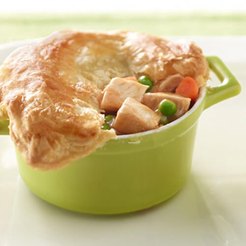 A flaky puff pastry baked with turkey breast, peas, carrots and gravy, served in green miniature bowls on a white platter.