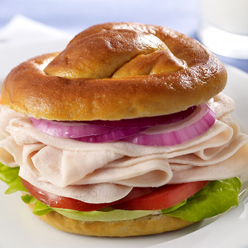 A hearty pretzel roll filled with lettuce, tomato, onion and turkey, served with mustard on a white plate with a side of carrots.