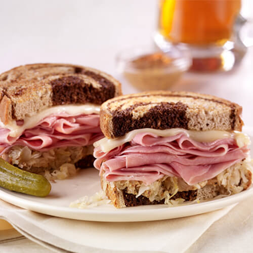 Turkey pastrami topped with sauerkraut and Swiss cheese on Pumpernickel on a white plate next to a pickle.