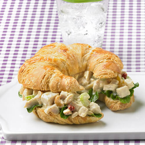A flaky croissant filled with turkey, vegetables, cashews, a mango chutney, smooth yogurt, and a curry mayo spread with a side of grapes, on a white platter on a purple checkered tablecloth.