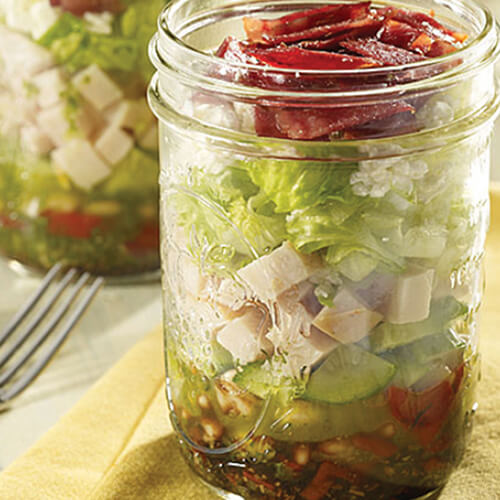 A hearty portion of balsamic vinegar, pesto, red pepper, beans, cucumber, turkey, feta, lettuce and bacon, served in a mason jar atop a yellow cloth.