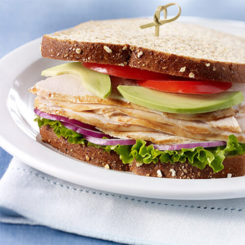 Whole grain bread layered with layer lettuce, onion, turkey, avocado and tomato, served on a white plate with potato chips, on a blue and white tablecloth.