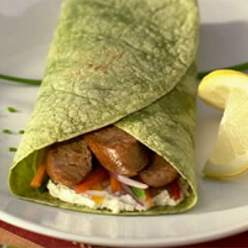 Fresh veggies, herbed goat cheese, sweet turkey sausage, and a tangy vinaigrette inside a spinach tortilla, on a white plate garnished with a lemon slice.