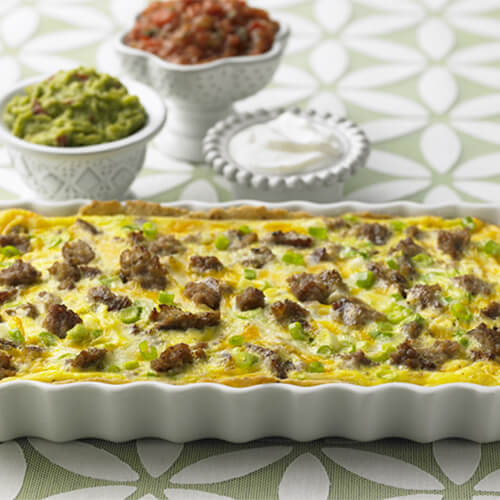 A hearty portion of eggs, turkey sausage, vegetables, and smoky chipotle adobo sauce baked over corn tortillas, served with guacamole, sour cream, and salsa, in a white baking dish, served on a green floral cloth.