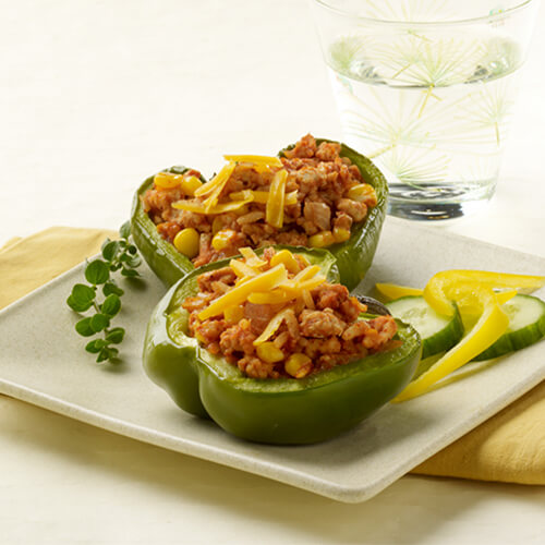 Colorful bell peppers filled with rice, corn, ground turkey, and topped with melted Cheddar, served with cucumbers and lemon on a white tray on a white table, served with a glass of water.