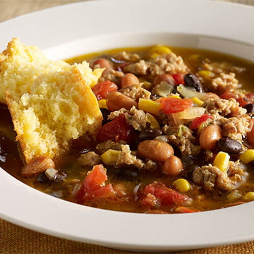 A hearty chili made with ground turkey, tomatoes, beans, and corn, served with corn muffins in a white bowl.