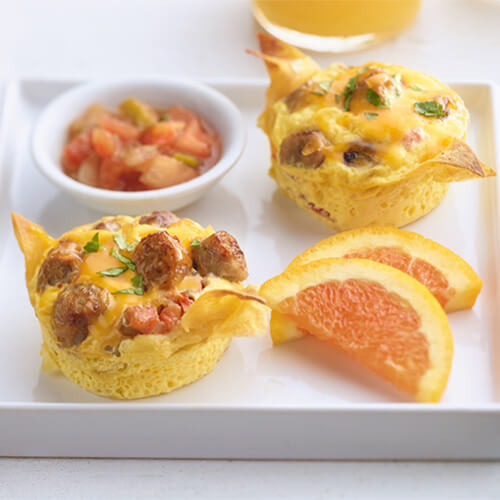 2 turkey taco cups baked with eggs, cheddar cheese, and lean turkey sausage baked with salsa and cilantro, served with a side of salsa and orange slices on a white plate and table.