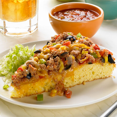 One hearty slice lean cornbread topped with ground turkey, fresh peppers and taco sauce on a white plate with a side of salsa.