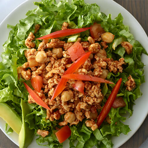 A substantial portion of lettuce, turkey, tomatoes, peppers, and cheese on a white plate, atop a wooden placemat with a linen tablecloth.