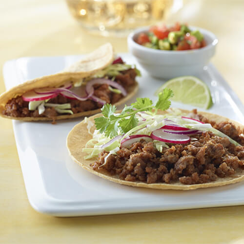 Turkey tacos filled with onions, radishes and cabbage, served with a side of homemade salsa, on a white plate atop a yellow tablecloth.