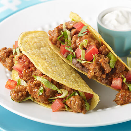 2 simple tacos filled with beans, ground turkey, tomatoes and lettuce, with a side of sour cream and lime wedges, served on a white plate on a cyan tablecloth.