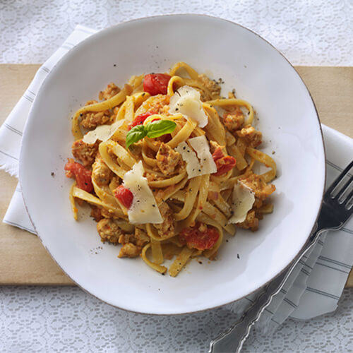 A hearty amount of fettuccine, mixed with lean turkey, garlic, white wine and pesto, sprinkled with shavings of parmesan cheese, served in a white plate on a wooden pizza board, on a linen tablecloth.