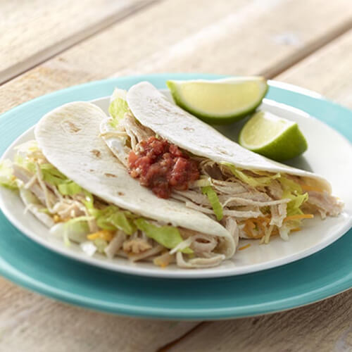 2 tortillas filled with lettuce, taco cheese, turkey breast, salsa, and guacamole, on a white plate garnished with lime atop a blue plate.