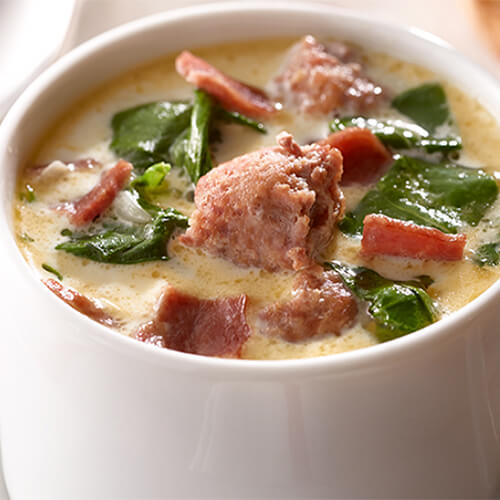 A creamy, meaty soup filled with spinach, potatoes, sausage and bacon, filled in a white bowl atop a white cloth.