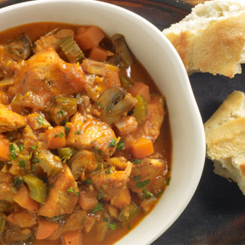 A savory soup, packed to the brim with veggies and turkey in a white bowl, served with torn bread on a wooden plate.