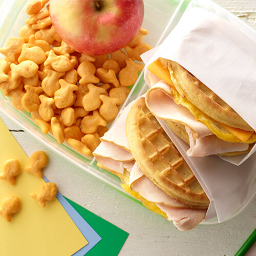 Turkey waffle sandwiches with golden fish crackers and an apple on a white plate.