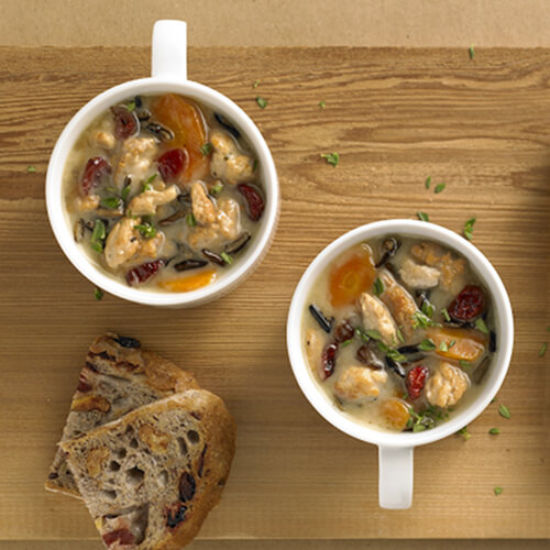 Two mugs filled with turkey, wild rice, and vegetables and Herb-Ox chicken bouillons with a side of slices of olive bread on a wooden platter with a yellow napkin on a tan background.