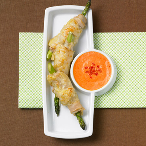 Stalks of asparagus wrapped in JENNIE-O® turkey breasts, served with a sauce made from and garnished with red bell peppers, on a white platter with a green napkin below on a brown background.