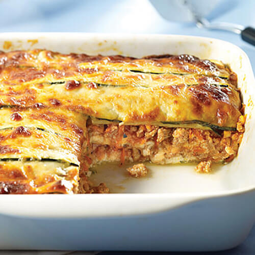 A hearty lasagna stuffed with basil, tomato sauce, zucchini, turkey, and mozzarella in a white lasagna pan, on a blue tablecloth.