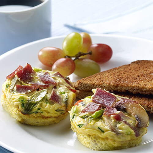 2 hearty frittatas chock full of vegetables, turkey bacon, and Swiss cheese, served on a white plate with a side of toast and grapes.