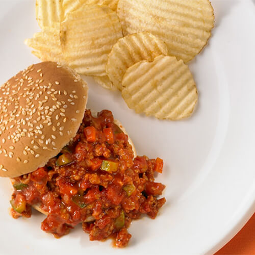 Turkey-based sloppy joes packed with vegetables such as zucchini, tomatoes, and carrots on a sesame seed bun, with a side of crinkle-cut potato chips served on a white plate, on a orange tablecloth.