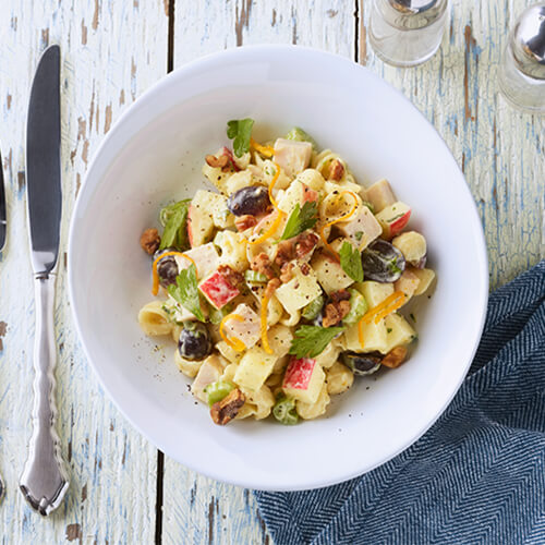 Pasta, fresh veggies and turkey all combined in a variation of a Waldorf salad, served in a white bowl, atop a linen cloth and wooden table.