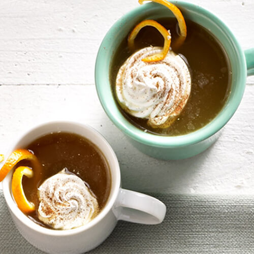 Warmed apple cider, spiced generously with cinnamon and topped with whipped cream and orange slices, served in mugs on a white table.