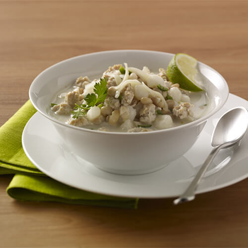 A chili made from white beans, hominy, spices, and ground turkey, combined and garnished with parsley and cheese, with a lime wedge, made in a white soup bowl and plate, on a green napkin, atop a wooden table.