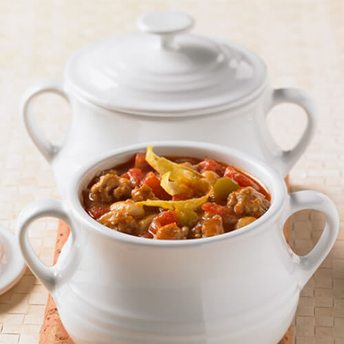 A healthy portion of turkey, tomatoes, beans, salsa, and tortilla strips in a white pot on an orange napkin on a white tablecloth.