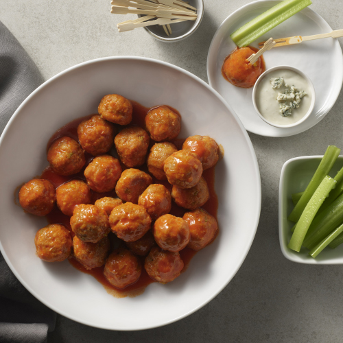 Spicy buffalo sauce engulfs a mound of chicken meatballs inside of a white ceramic plate, while a side of cut celery and bleu cheese sauce accompanies on their own white dishes.